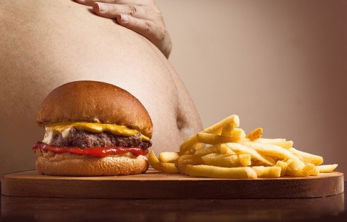 Foods that cause cancer: Image of a hamburger and fries on a table with a man's fat stomach in the background.