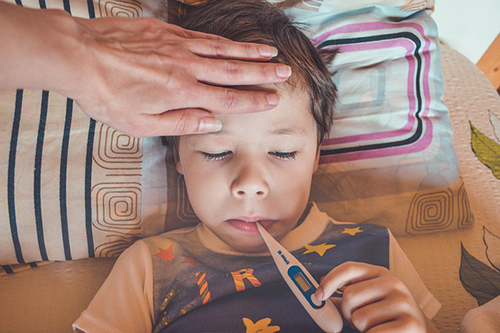 little boy with a thermometer in his mouth with mother's hand on his forehead making sure he does not have a fever