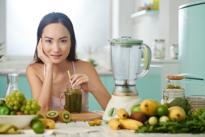 woman sitting at table with blender and fruits along with a blender and smoothie in a jar