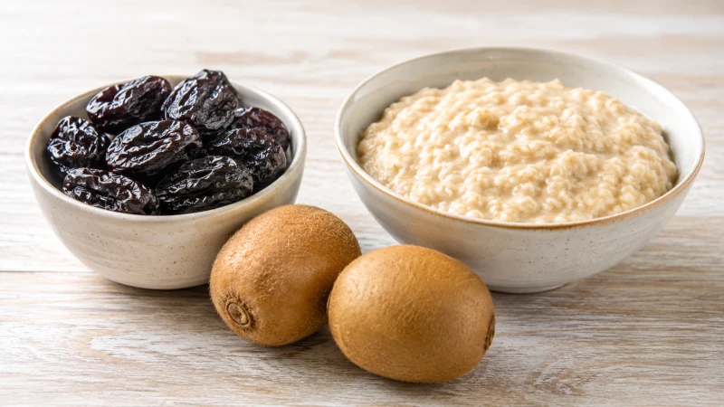 Bowl of prunes, two kiwifruit, and a bowl of oatmeal arranged together — the three most evidence-backed foods for constipation.