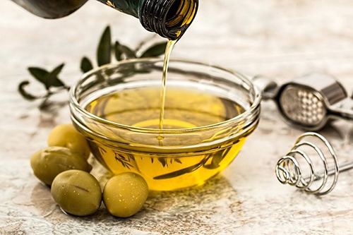 olive oil being poured into a glass container with olives around the container on a table
