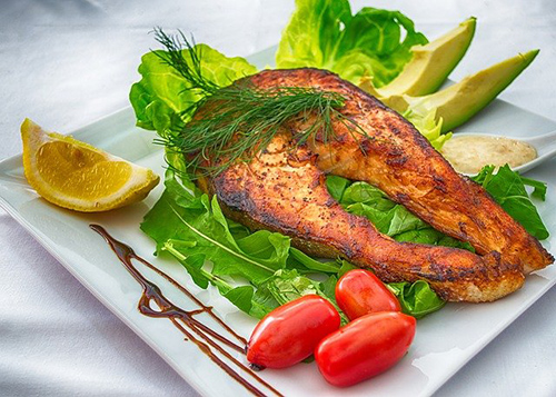 grilled salmon on a plate surrounded by lettuce, avocado, tomatoes, and garnished with a lemon
