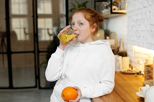 woman in white sweatshirt drinking a glass of orange juice while holding an orange in her hand