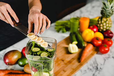 hands putting sliced bananas into a blender with other vegetables and fruits