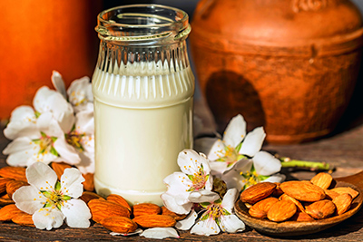 glass jar of almond milk on a table along with almonds and blossom surrounding the jar.