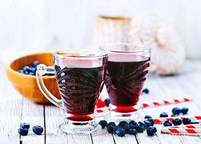 two glasses of blueberry juice with some blueberries scattered around a table