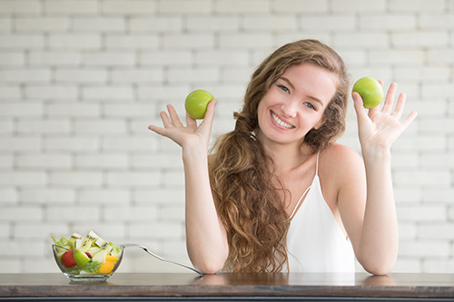 woman holding up two apples and a bowl of various fruits on the table for foods and health